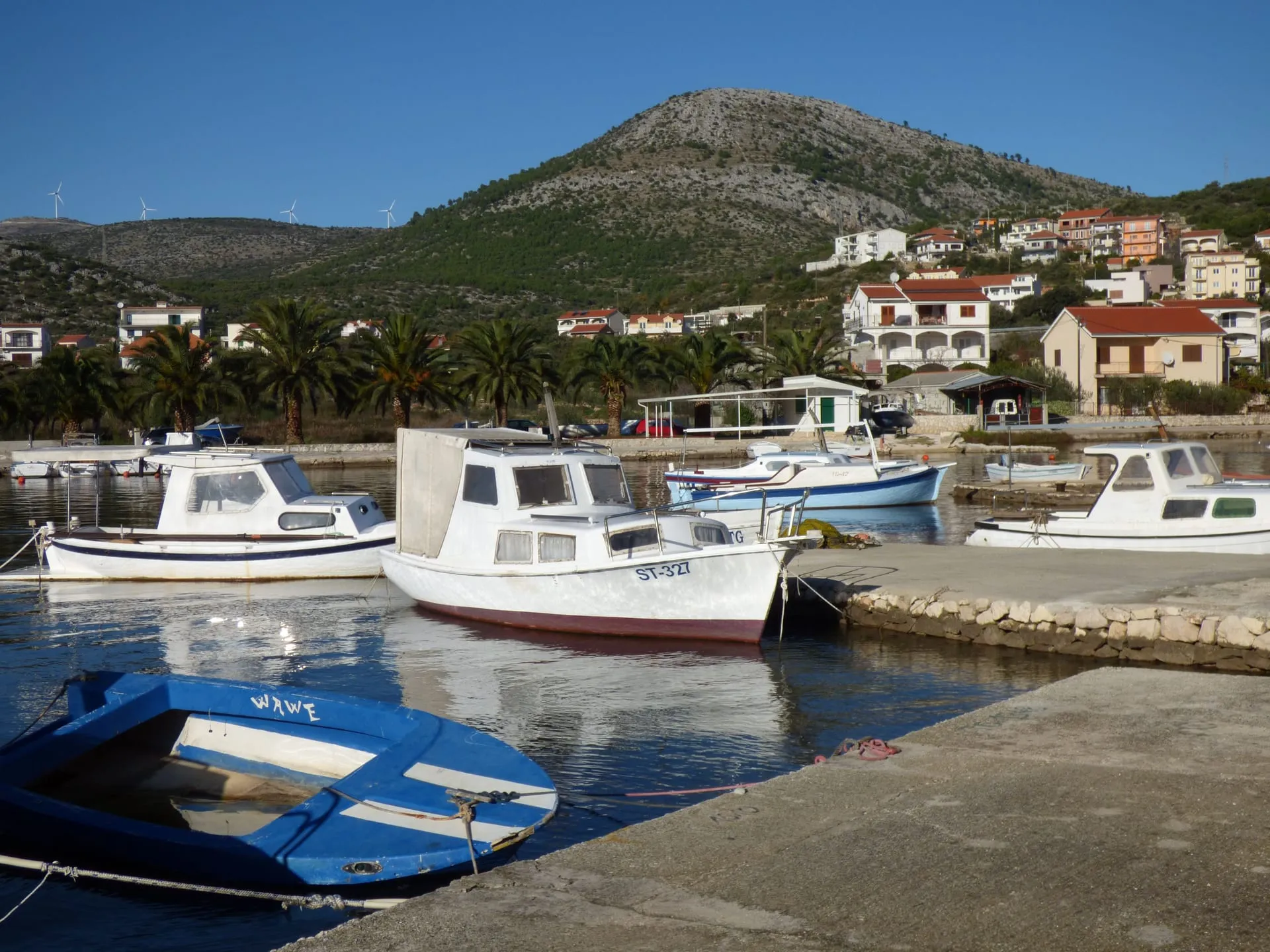 Small boats moored in Grebaštica harbour, with palm trees, houses and a green hill behind