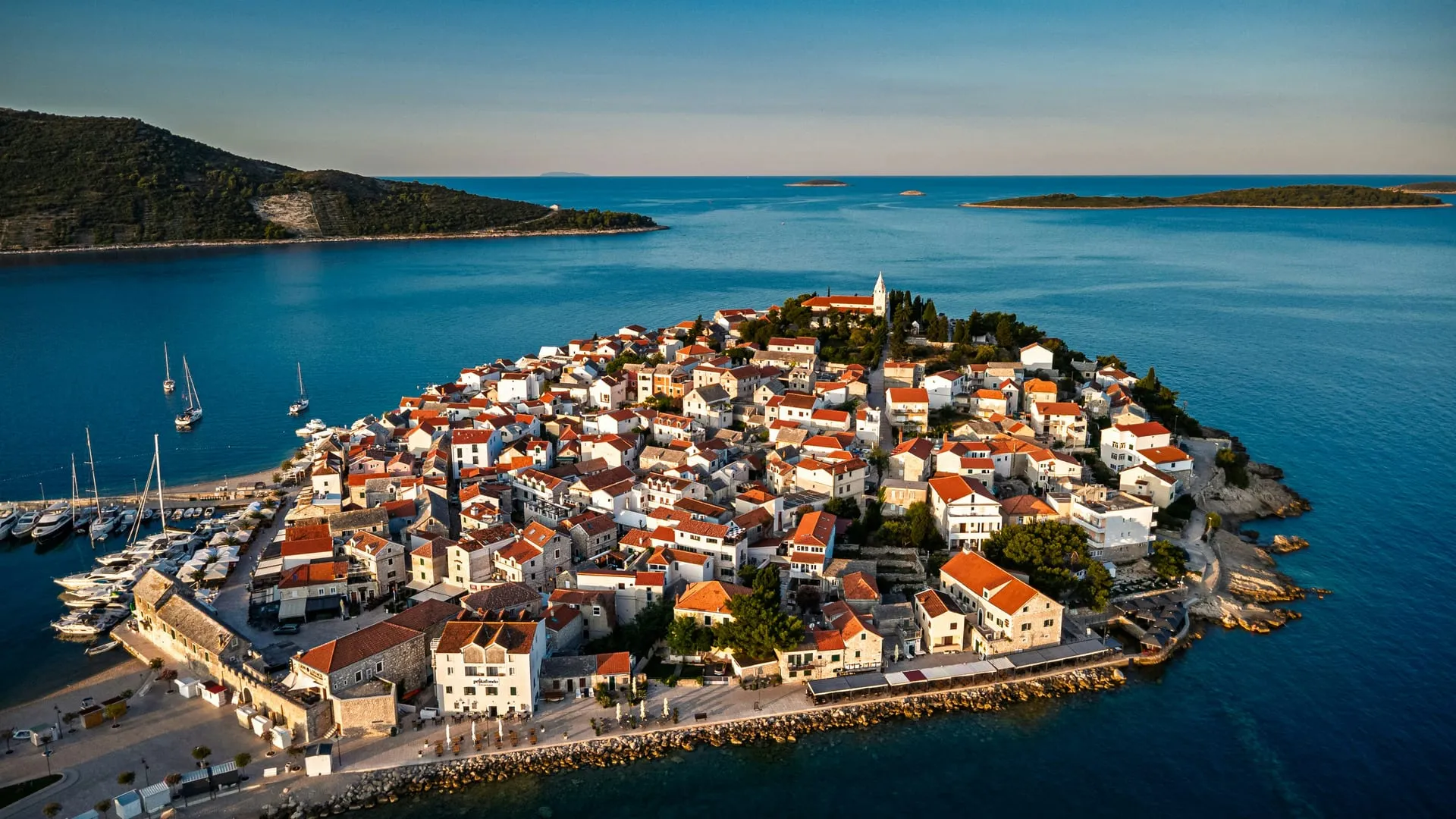 Primošten from the air: the old town's red roofs and church bell tower on a point surrounded by sea, sailing boats moored along the harbour