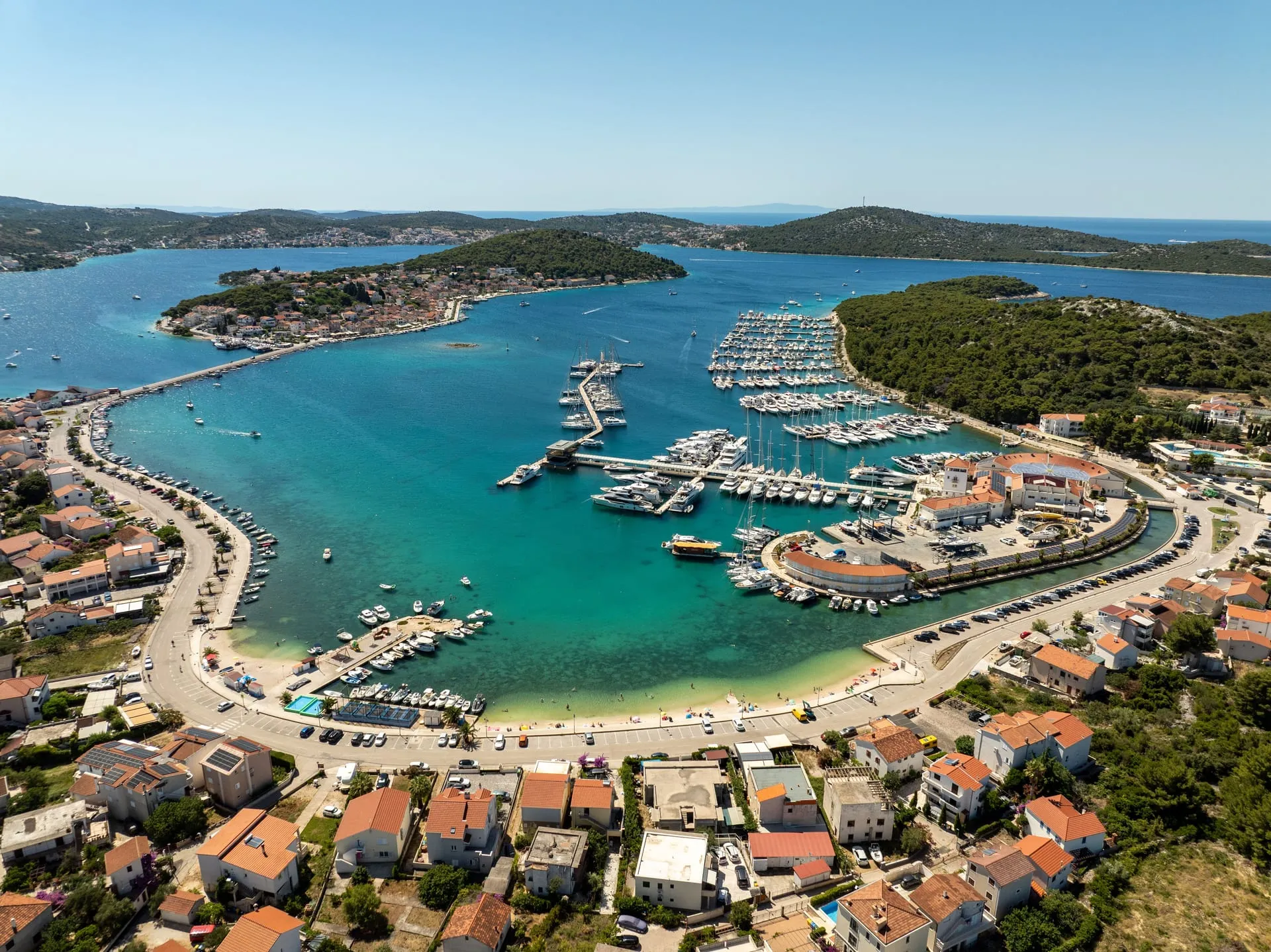 Rogoznica from the air: a turquoise bay with a marina and a beach, and the old town on an islet joined by a causeway